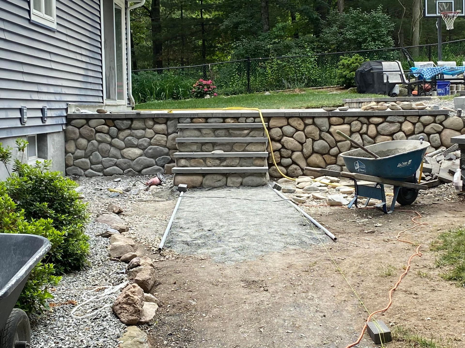 Backyard construction site: stone retaining wall, stairs, gravel pathway, wheelbarrow.