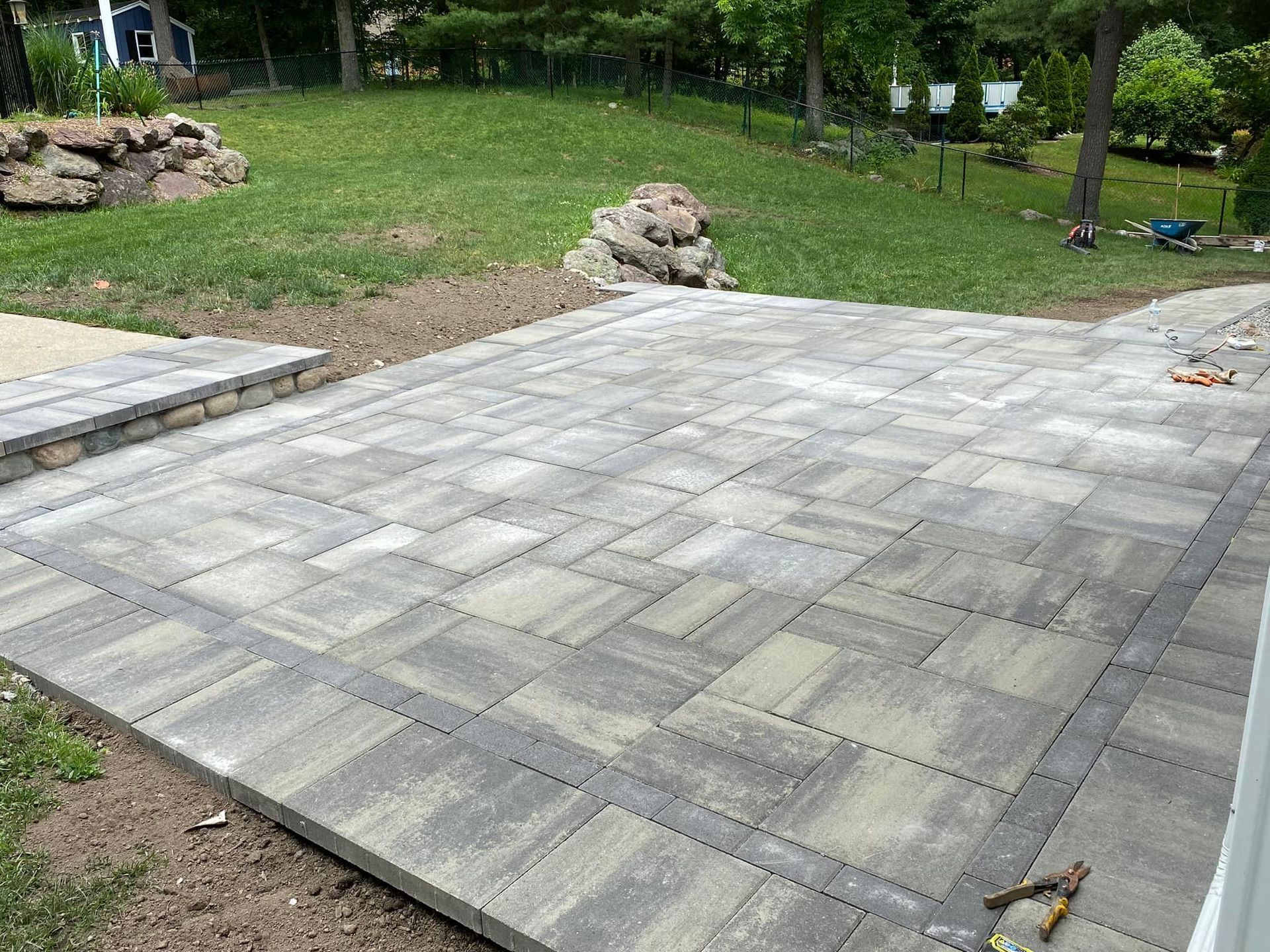 Stone patio in backyard, surrounded by grass and small retaining wall.