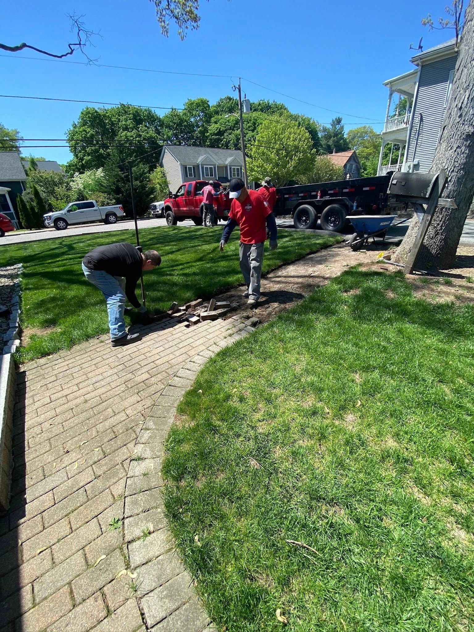Men work on a brick pathway next to grass. A truck and trees are in the background on a sunny day.