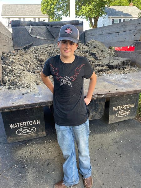 Boy in a baseball cap stands by a dump truck filled with asphalt, hands in pockets.