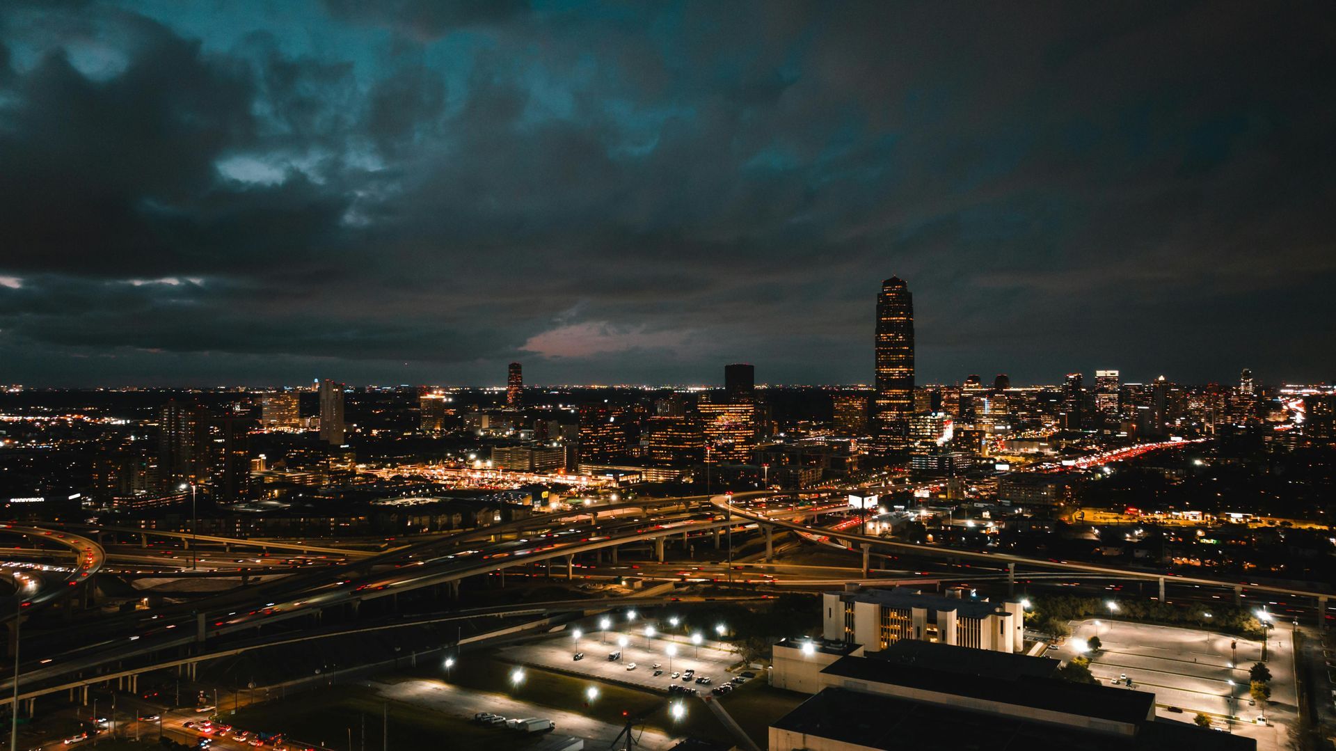 City skyline at dusk with glowing lights under a dark, cloudy sky