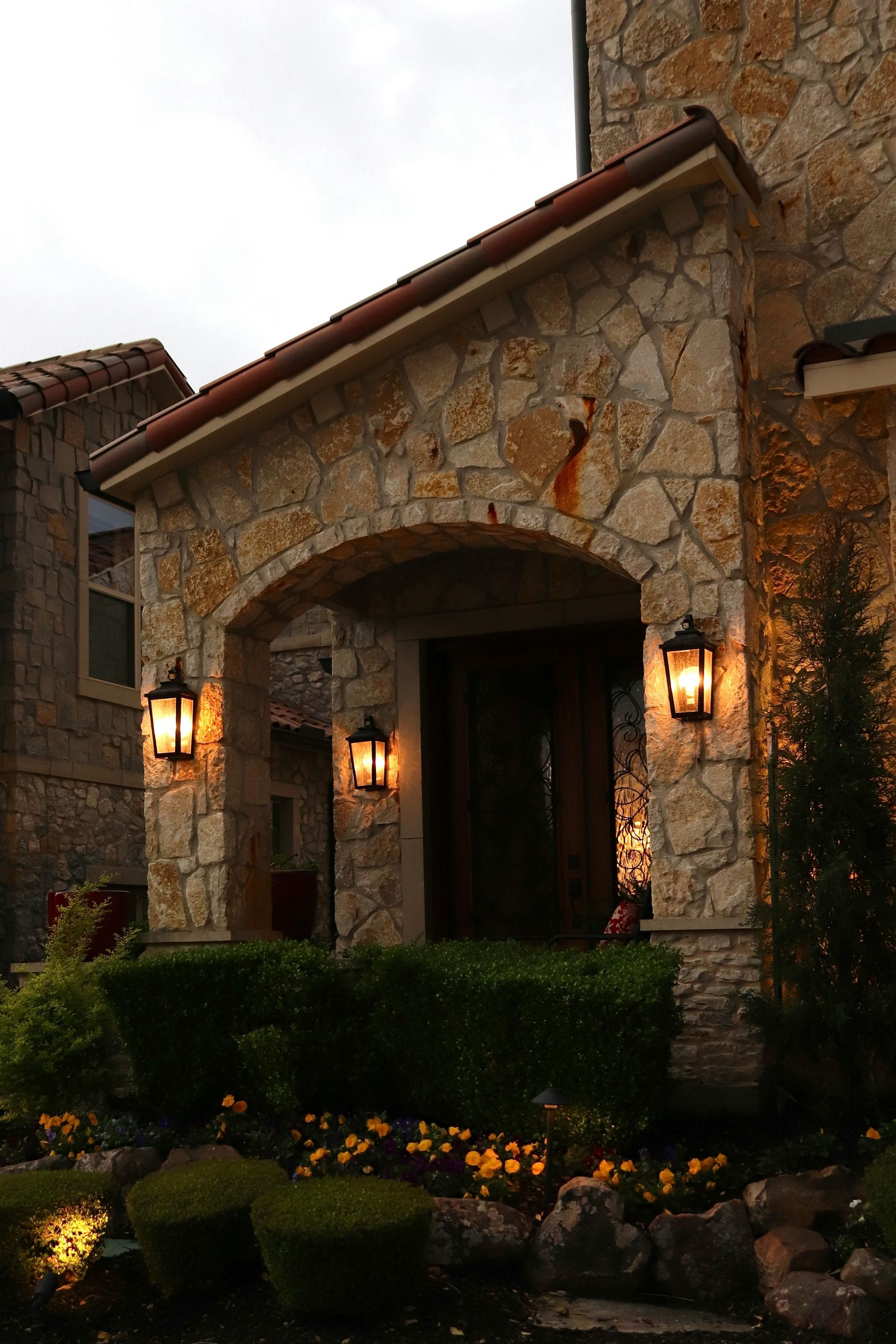 Stone house entrance with warm lanterns, arched doorway, and trimmed hedges at dusk