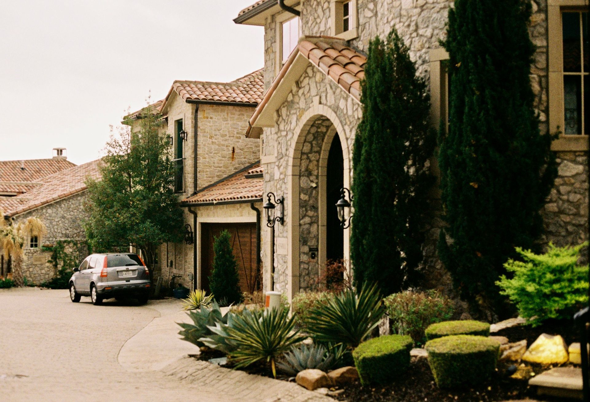 Stone house entryway with arched doorway, parked car, and landscaped greenery on a quiet driveway