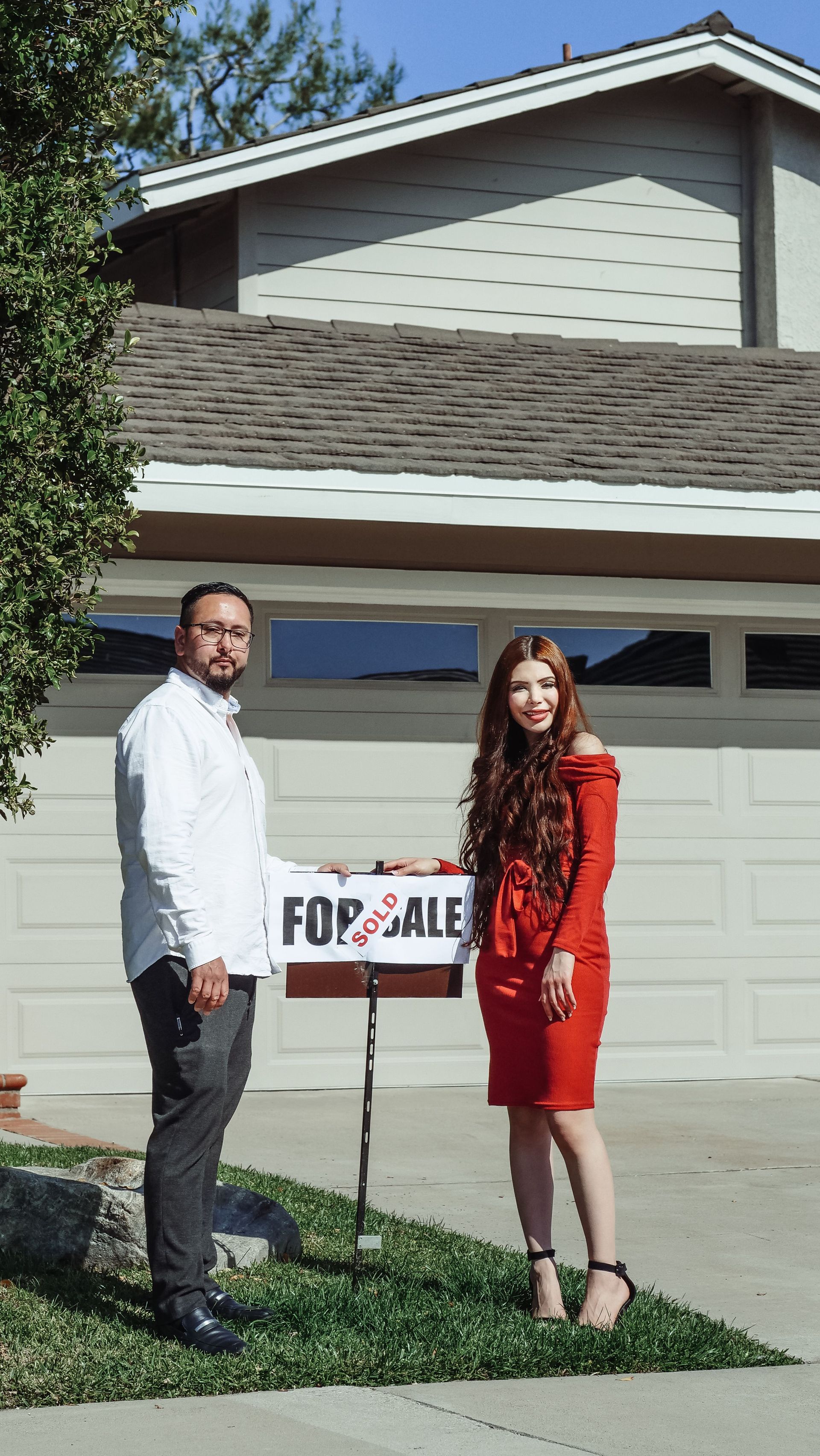 Two people stand outside a house holding a “For Sale” sign, with the garage in the background.