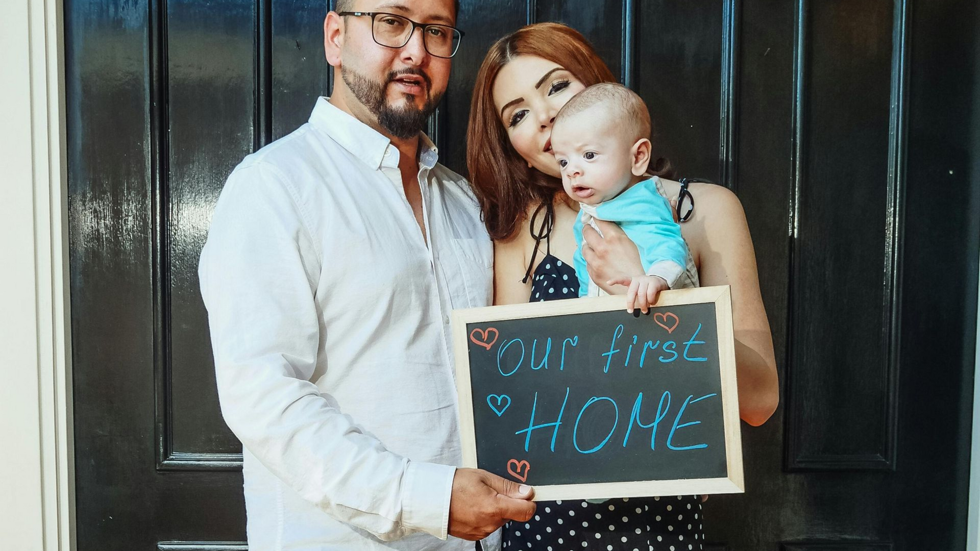 Family holding a cardboard sign that says “Our First Home” in front of a dark door.