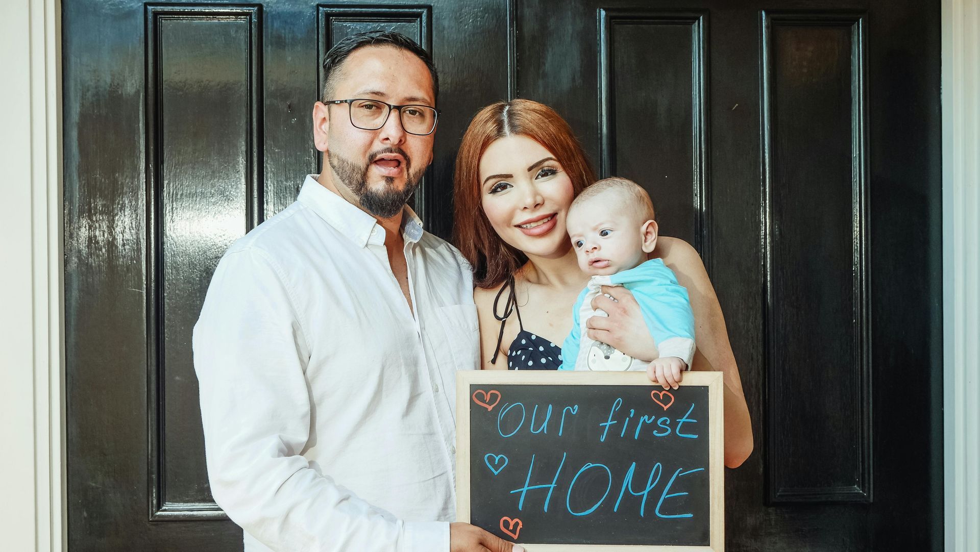 Family of three stands in front of a black door, holding a chalkboard sign reading