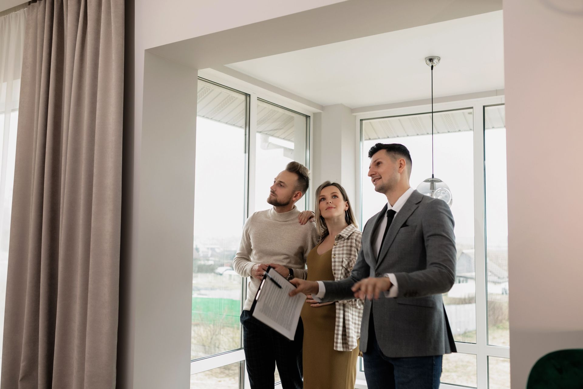 Three people discussing a property inside a bright, empty room with large windows.