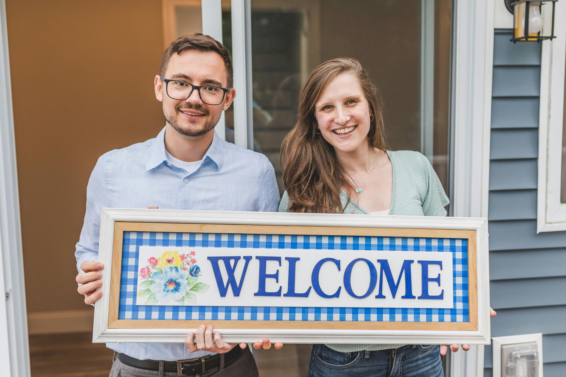 Two people holding a colorful “WELCOME” sign on a house porch