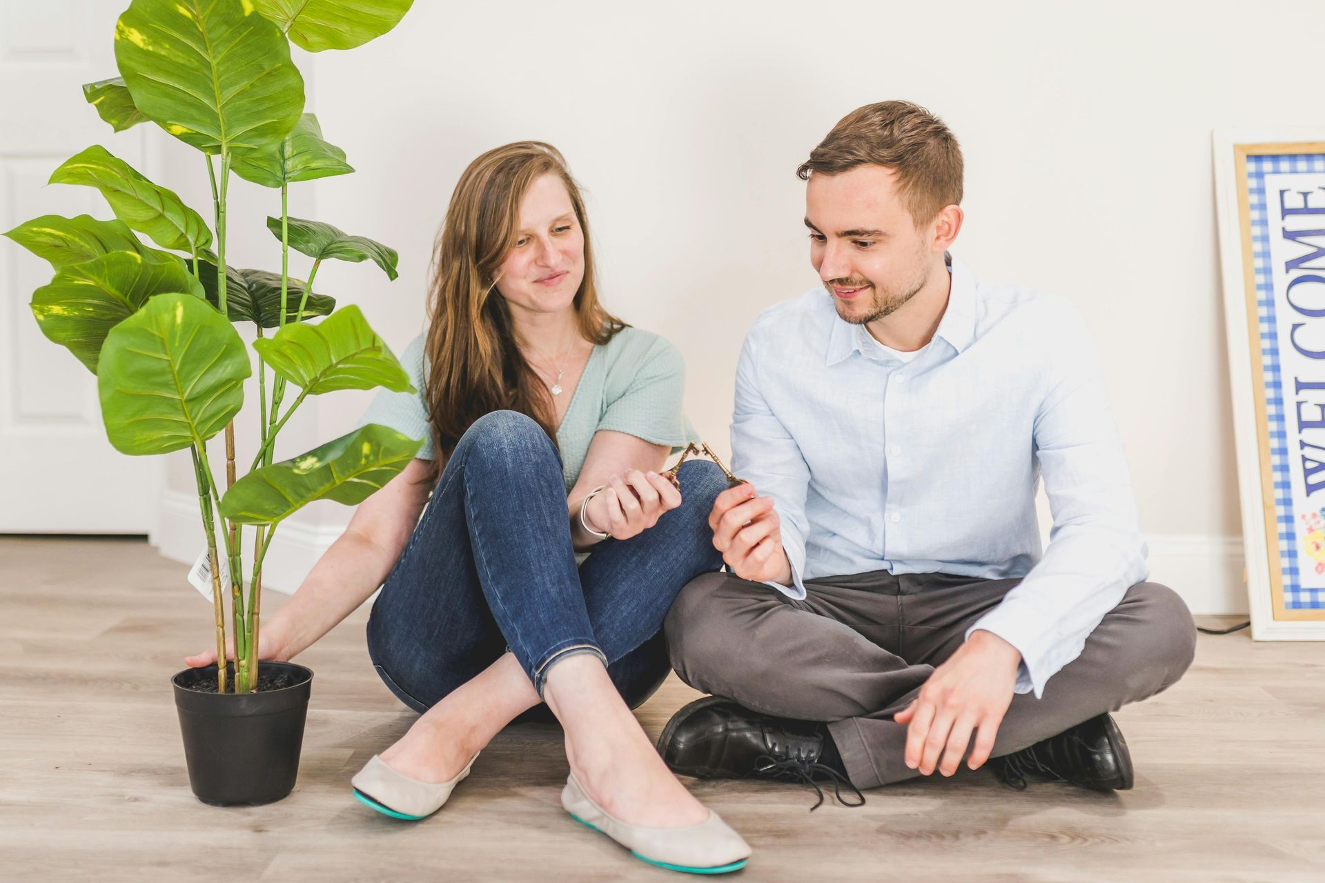 Couple sitting on floor, plant between them; woman in jeans, man in button-down shirt, both smiling.