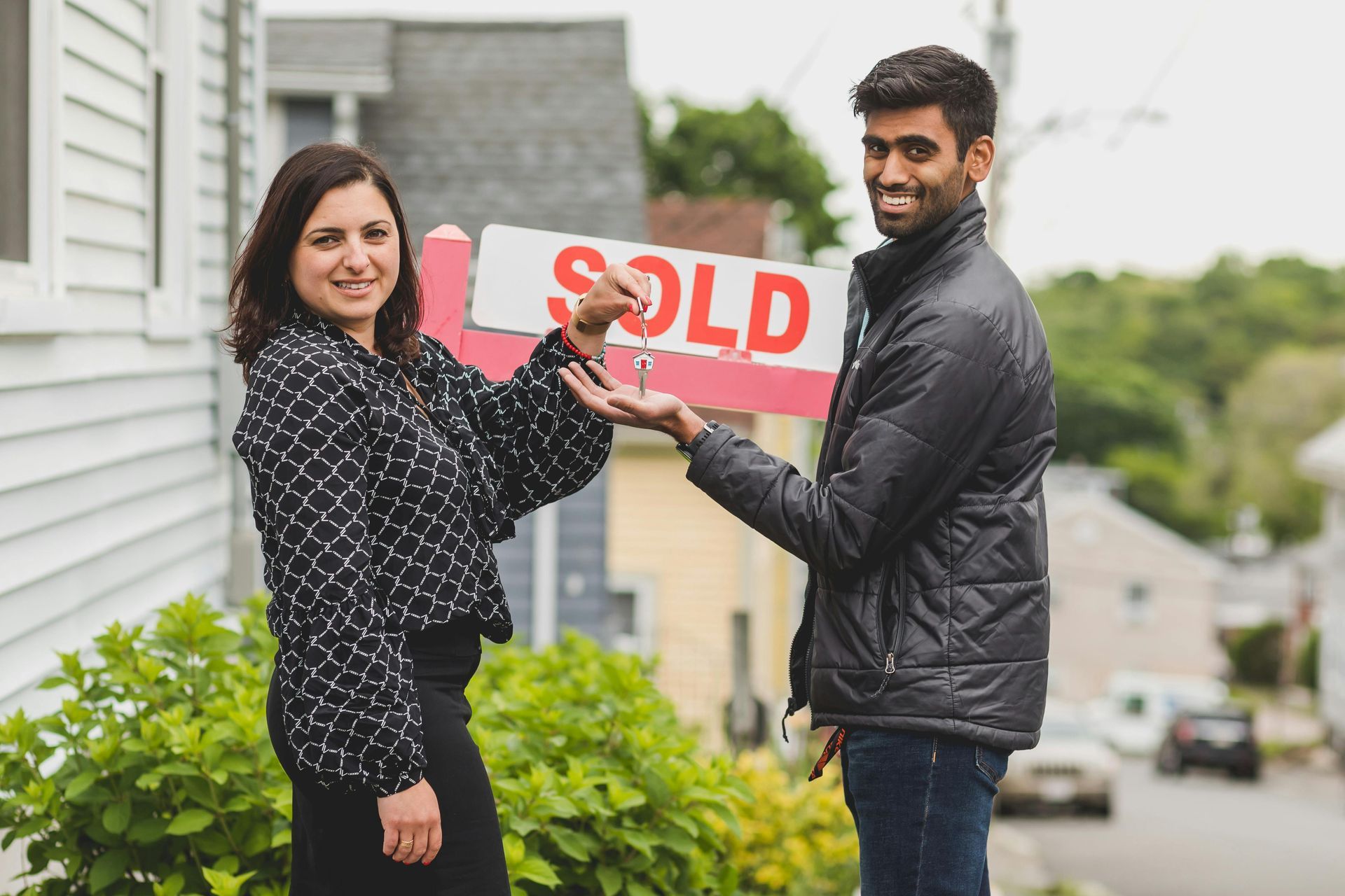 Real estate agent handing keys to a person in front of a house with a 