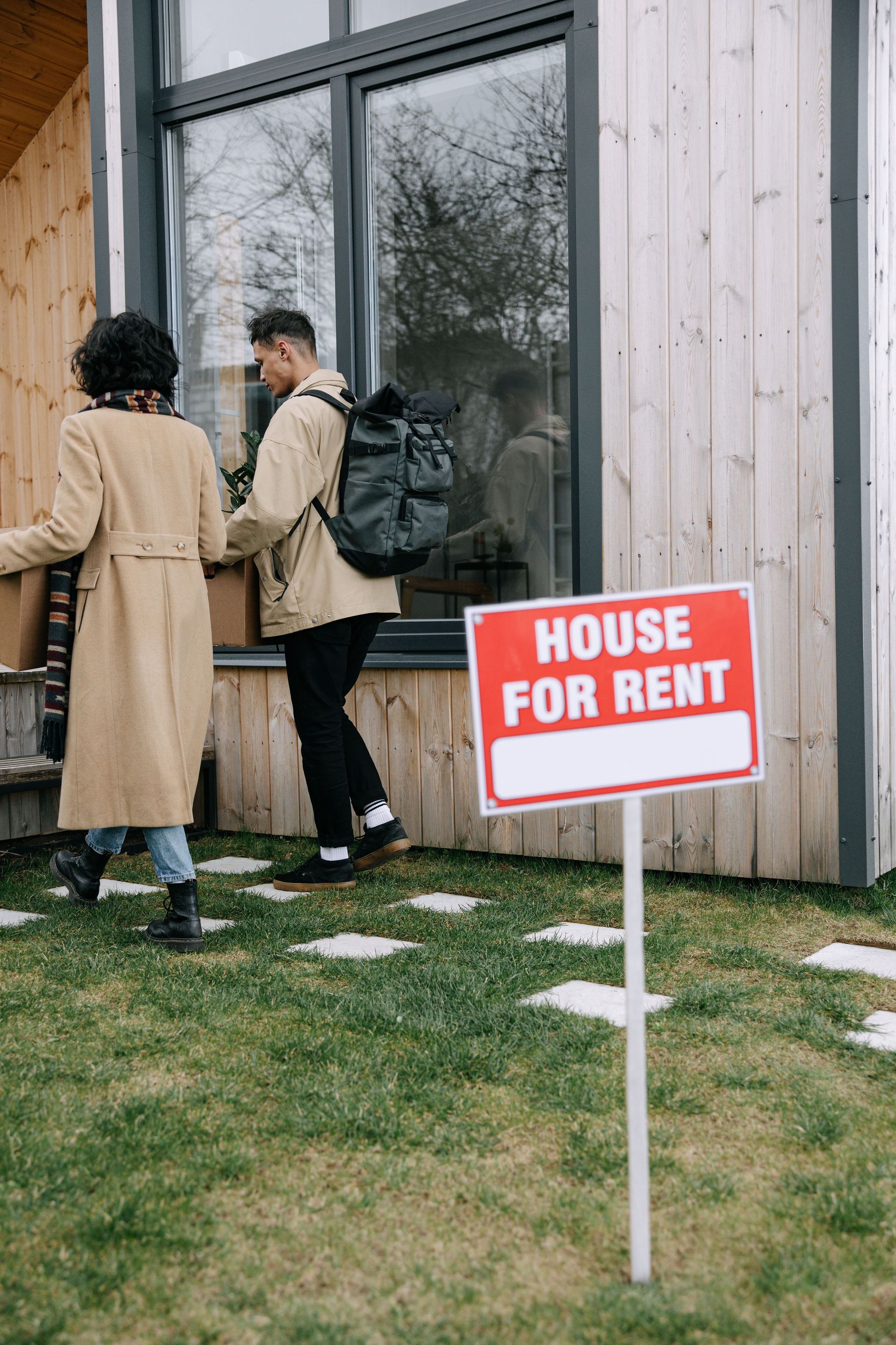 Two people outside a wooden house beside a red “HOUSE FOR RENT” sign.
