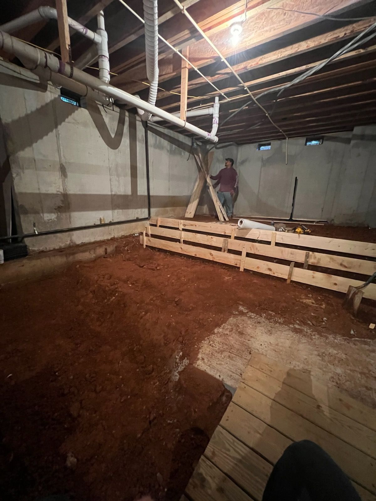 Basement under construction with wooden forms, red dirt floor, and person standing near a support.