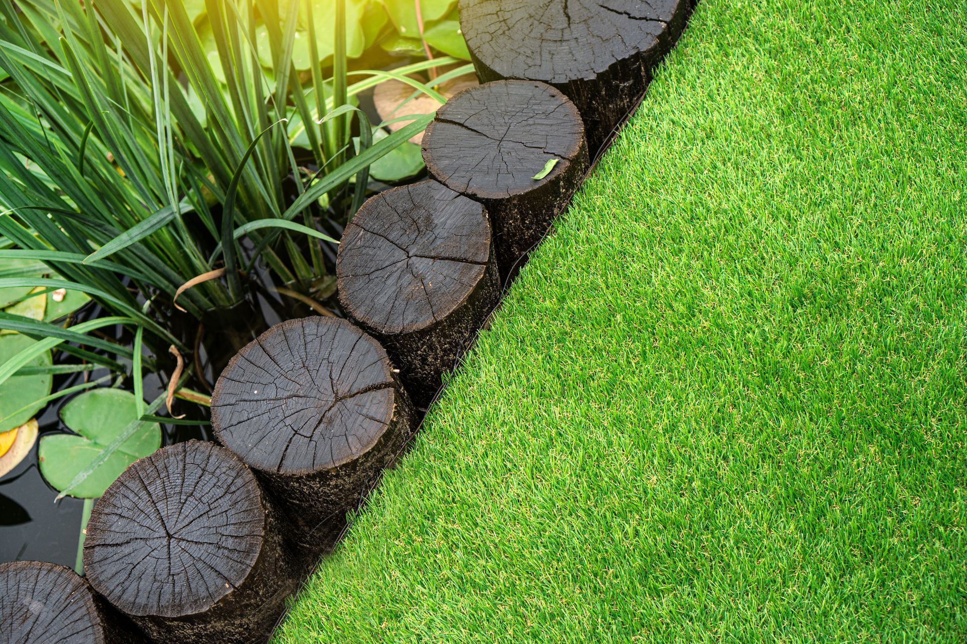 Wooden log path beside a patch of bright green grass and a water garden with greenery.