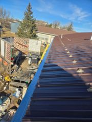 A view from a brown metal roof looking down at a cluttered yard with a lattice fence and a bright yellow ladder.