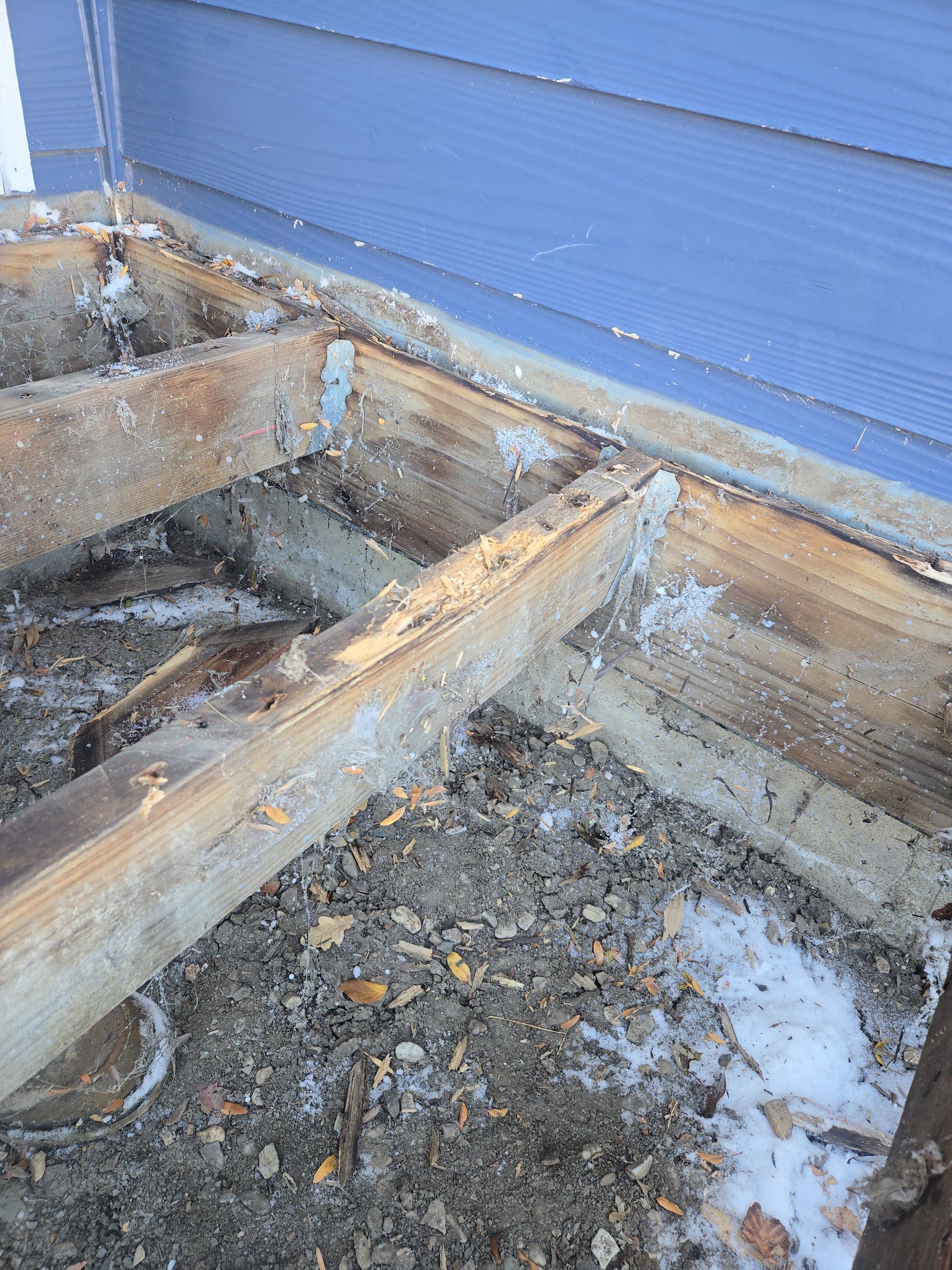 A close-up view of a deteriorating wooden deck frame attached to a blue house, with debris and snow on the ground below.