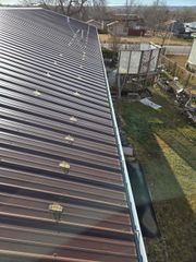 A high-angle view of a brown corrugated metal roof with solar heating coils, featuring a metal gutter and yard below.