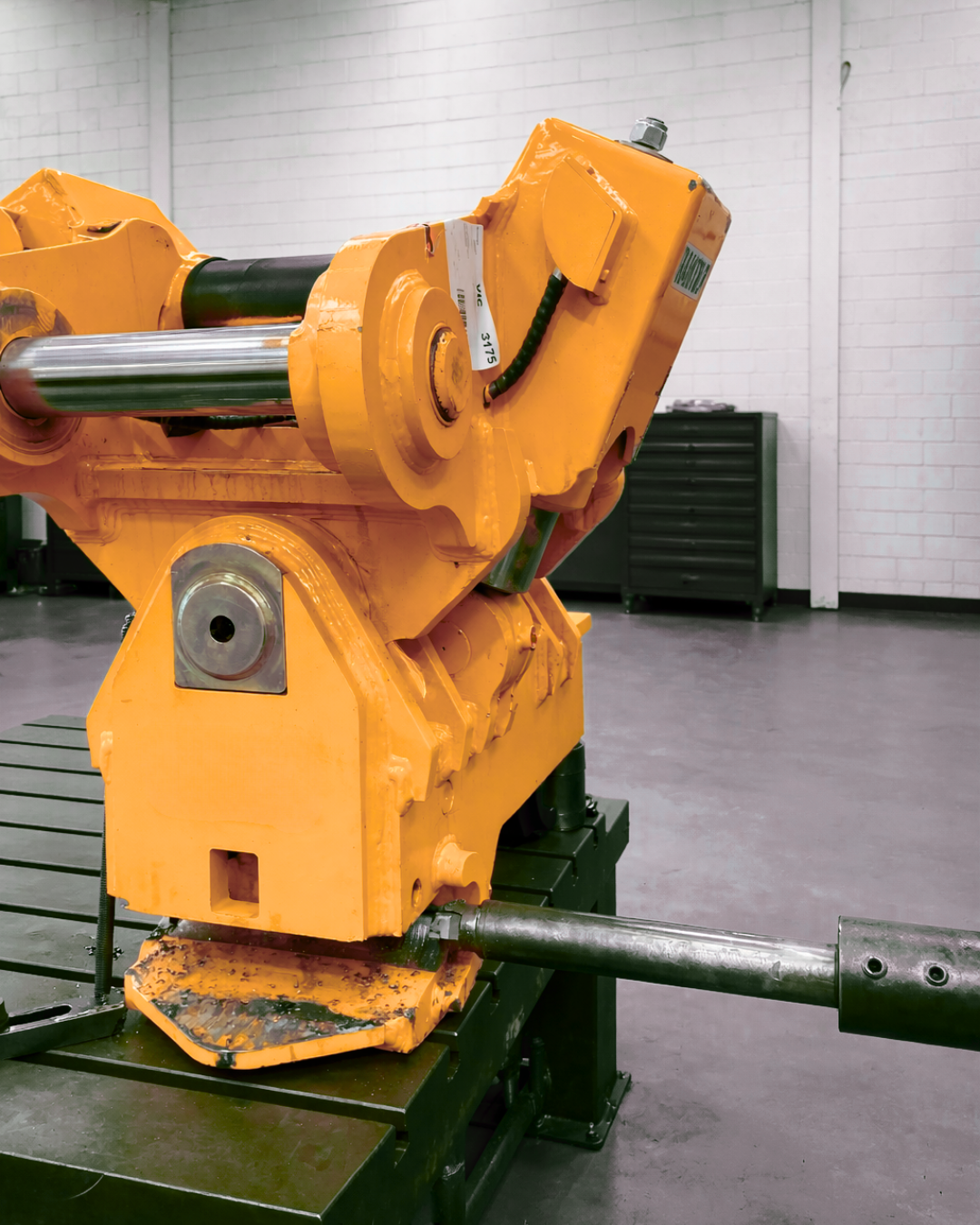 Yellow industrial machine in a warehouse, with a metal rod attached and green pallets in the background