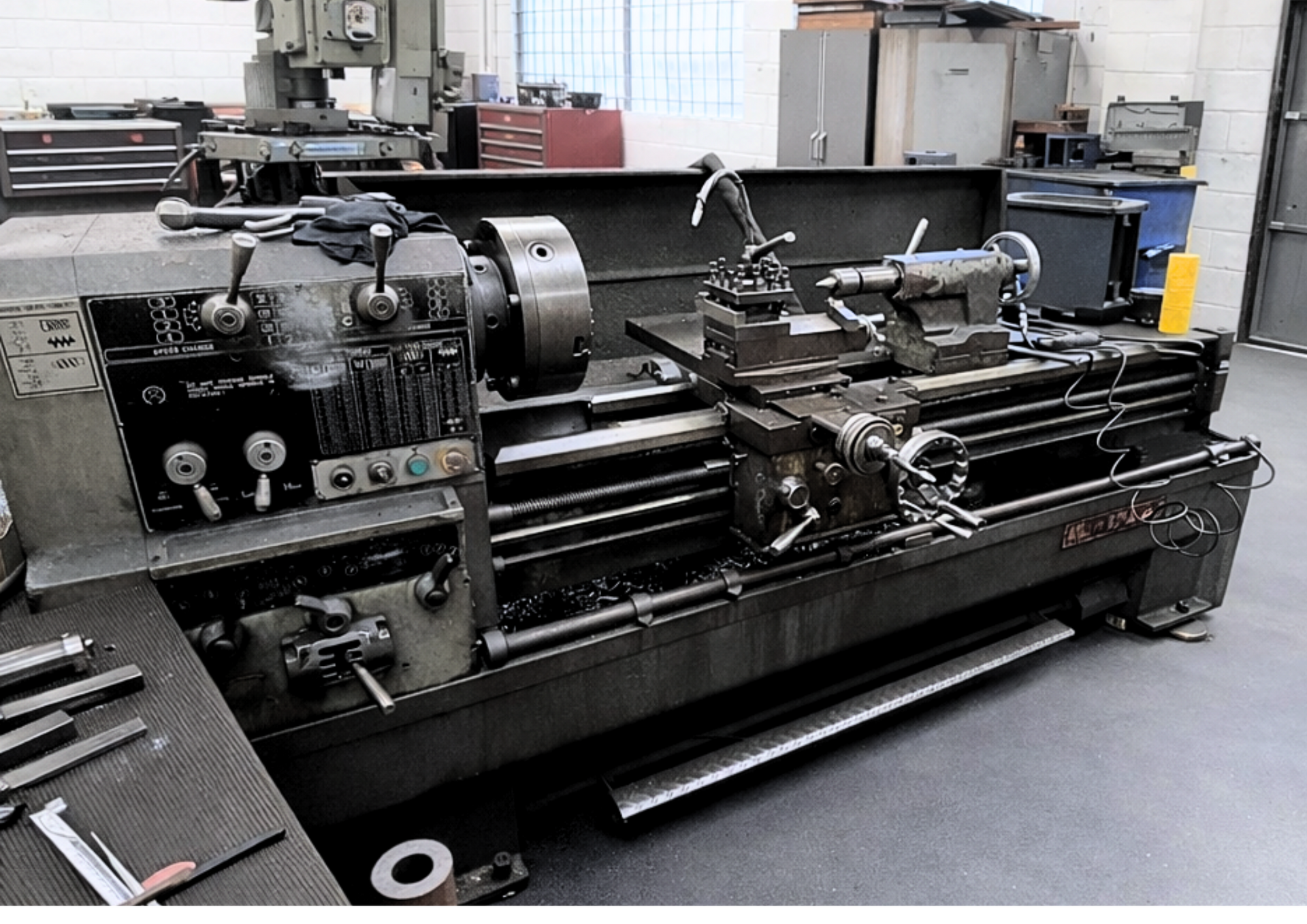 Large metal lathe in a machine shop with gray walls, windows, and industrial equipment.