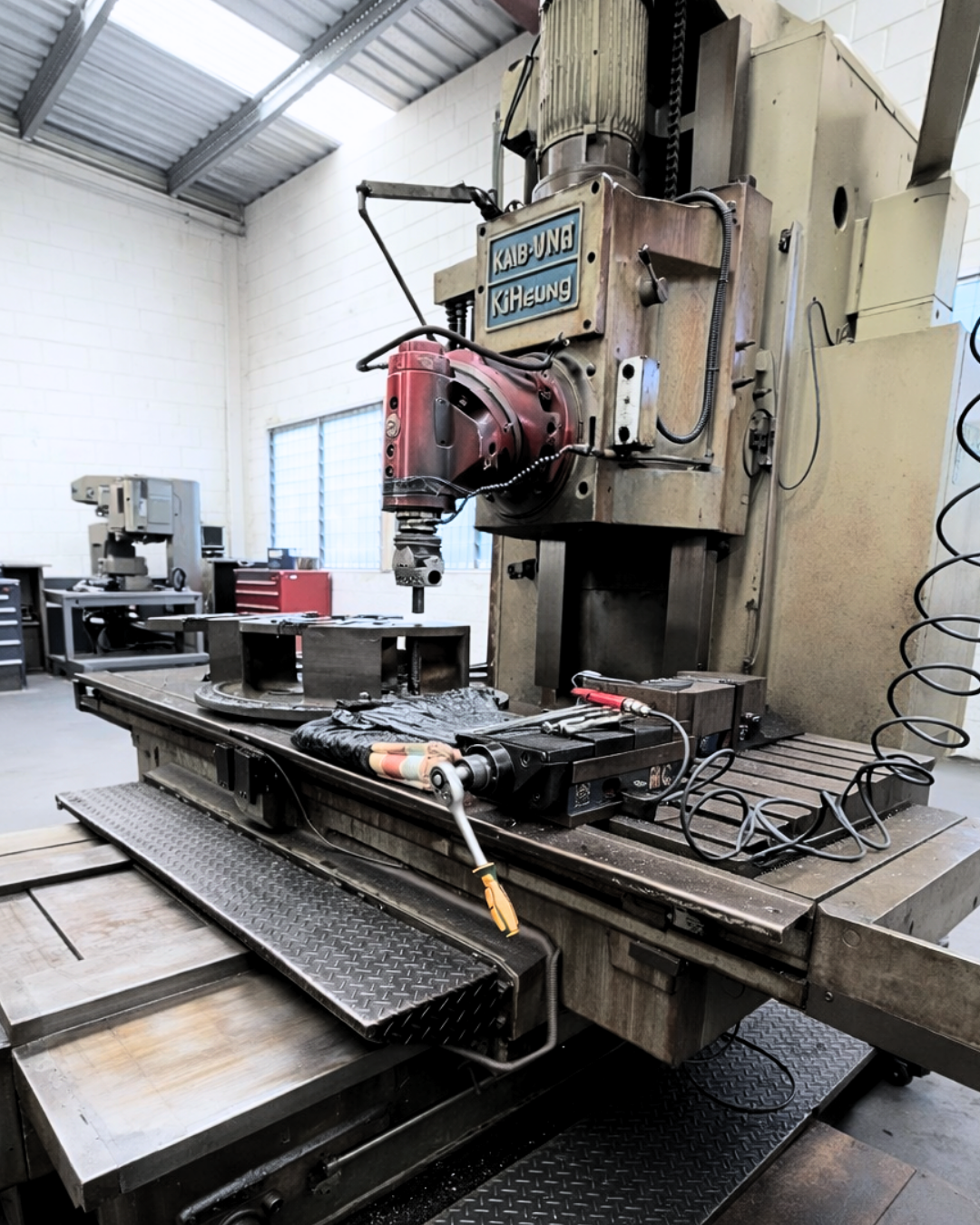 Industrial workshop with a large vertical milling machine and metal parts on a workbench.