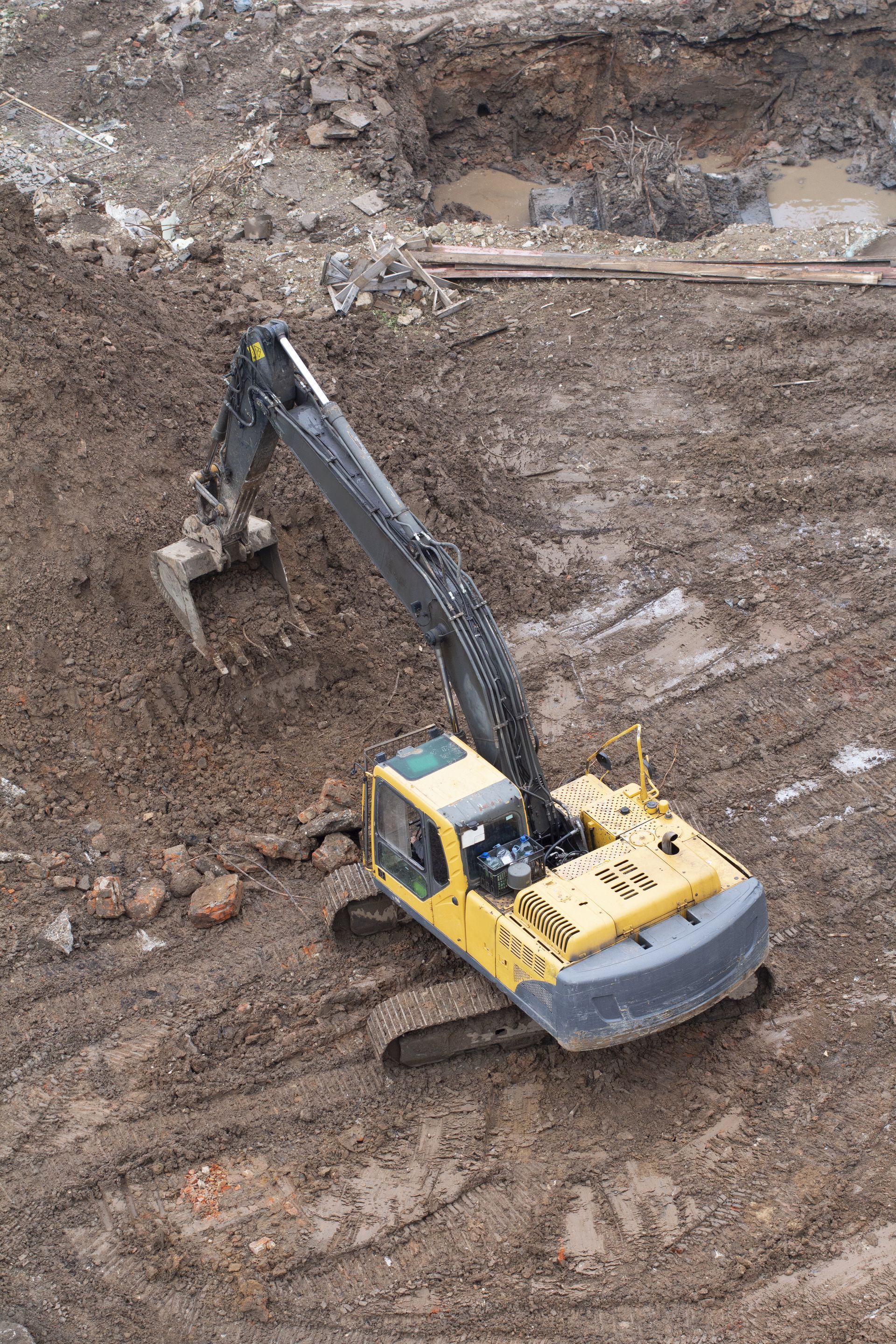A yellow tracked excavator with an extended arm sits on a muddy construction site, working the earth.