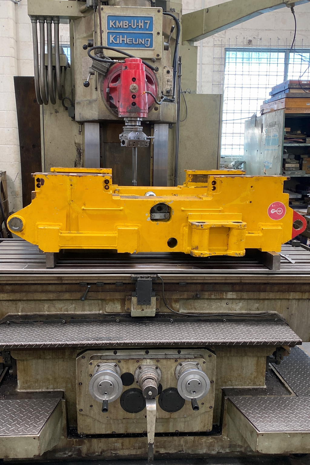 Yellow industrial machine part on a metal worktable inside a workshop, with a drill press behind it.