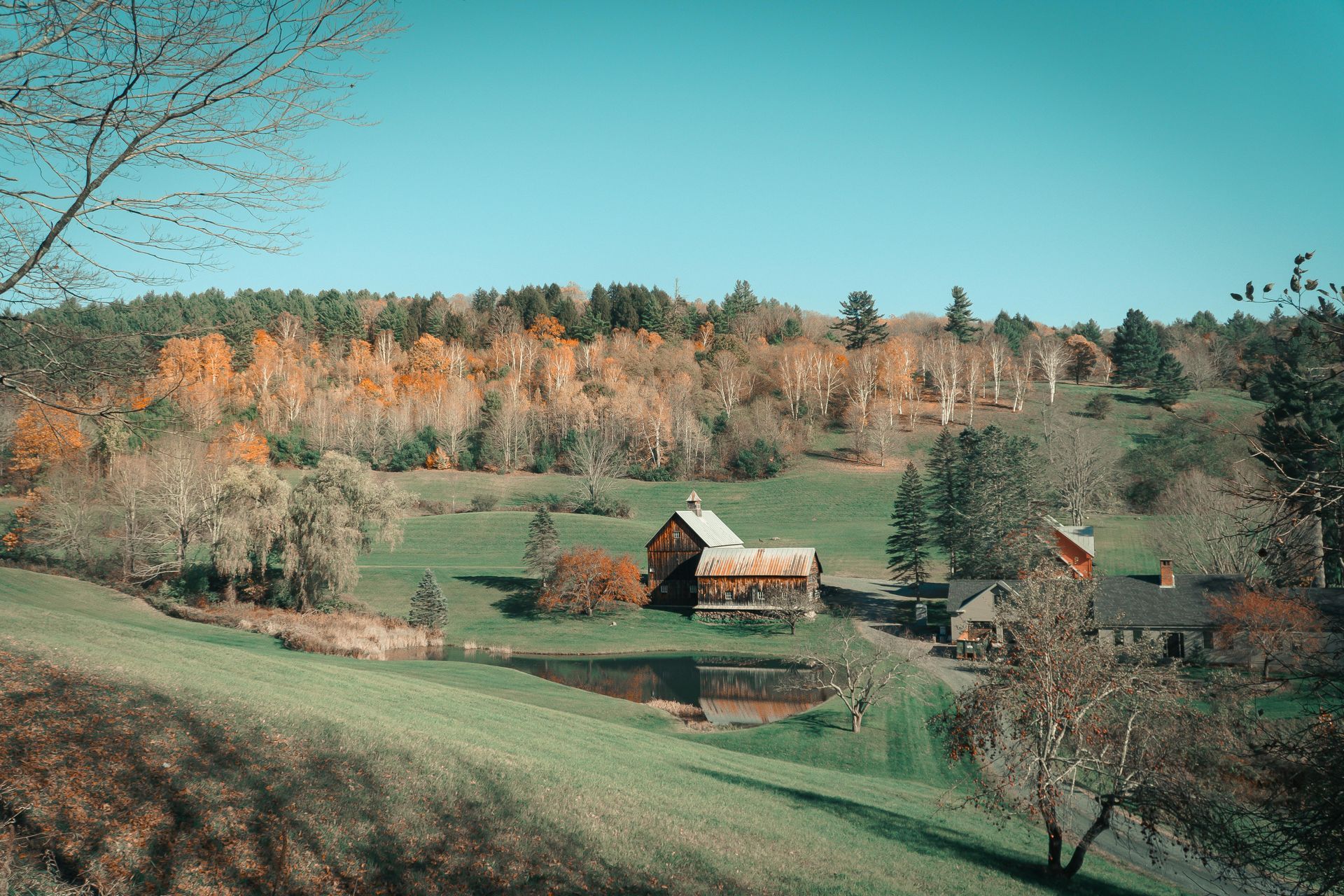 Rural valley with green fields, two small barns, and autumn trees under a clear blue sky