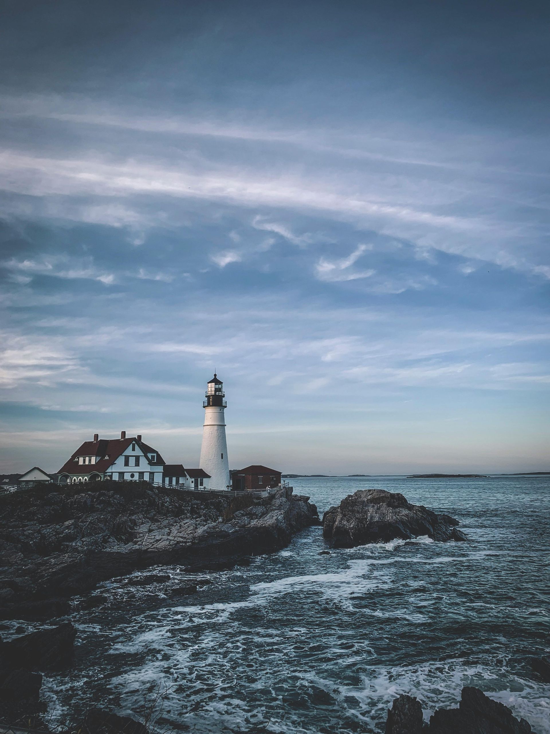 Lighthouse beside a coastal house on rocky shore under a cloudy blue sky