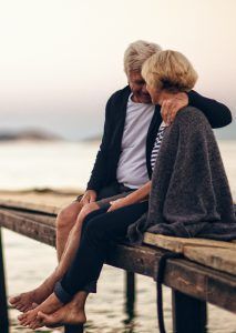 Couple sits closely on a wooden pier, embracing, overlooking the water at dusk.