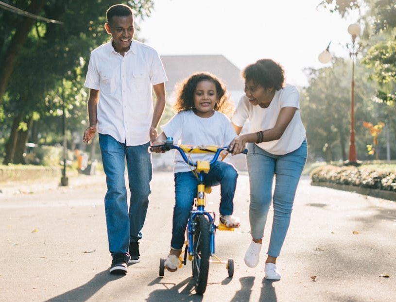 Family helps child ride a tricycle in a park.