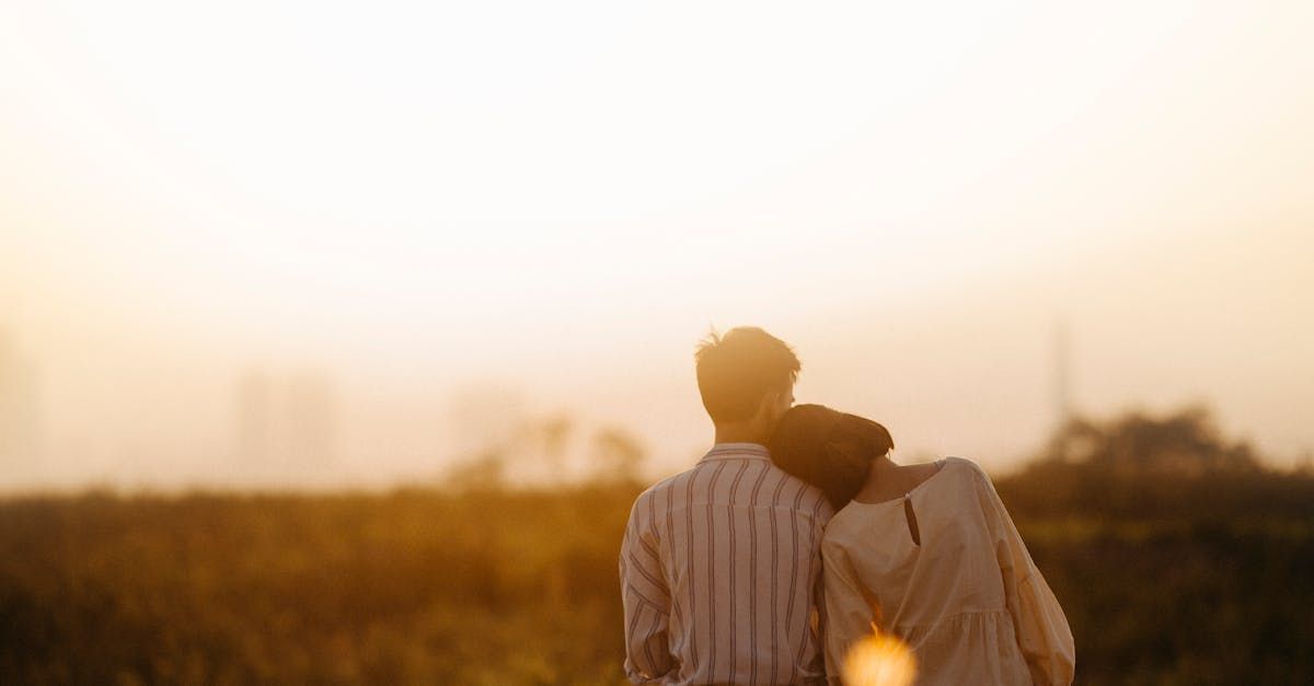 Couple standing in field, one resting head on the other's shoulder, facing a golden sunrise.