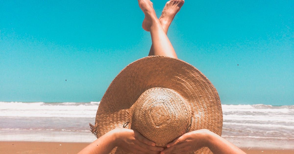 Person on beach, wearing large straw hat, legs up, blue sky and ocean in background.