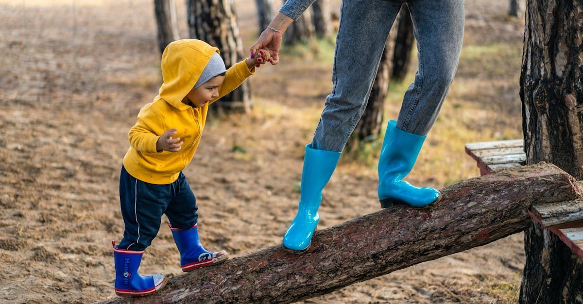 Child in yellow hoodie and blue boots holds adult's hand, walking on a wooden beam in a forest.