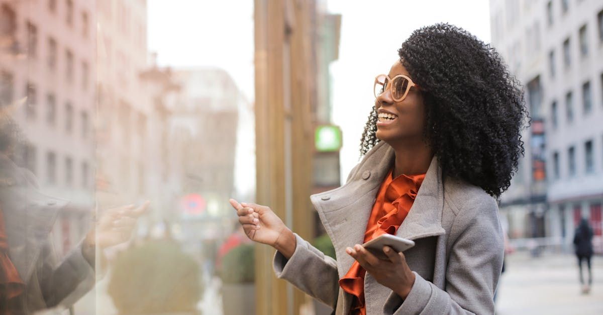 Woman looking in a shop window, smiling, wearing glasses and a gray coat, holding a phone, city background.