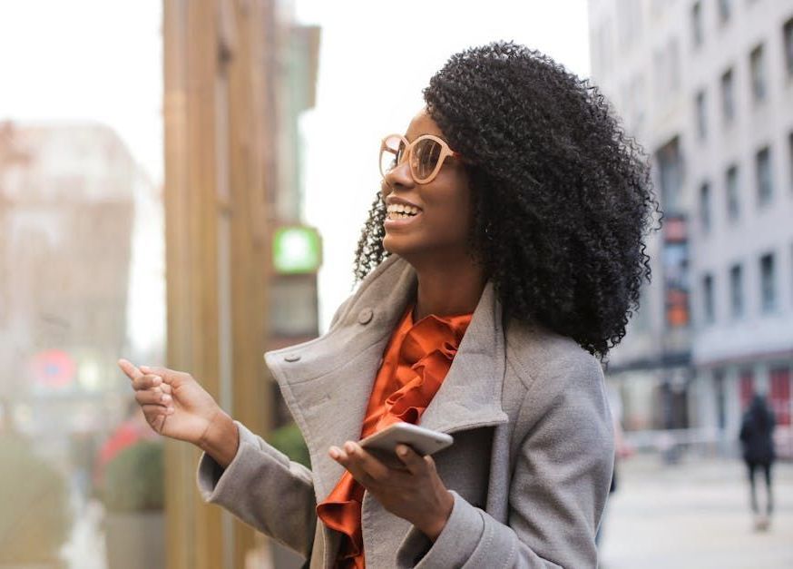 Woman with curly hair in a gray coat and glasses smiles, holding a phone, on a city street.