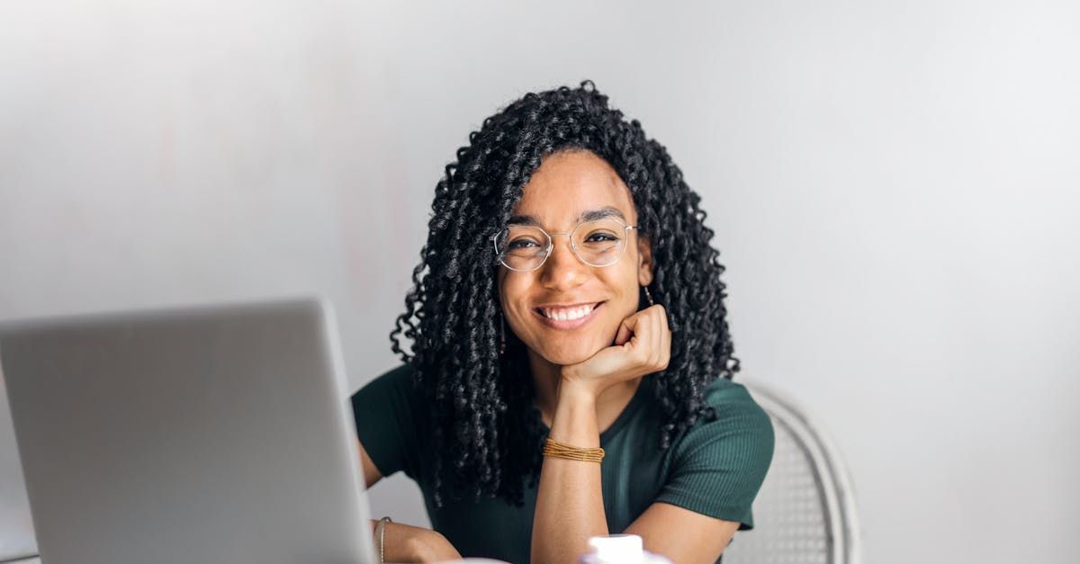 Woman with glasses smiles, resting chin on hand, seated at a laptop.
