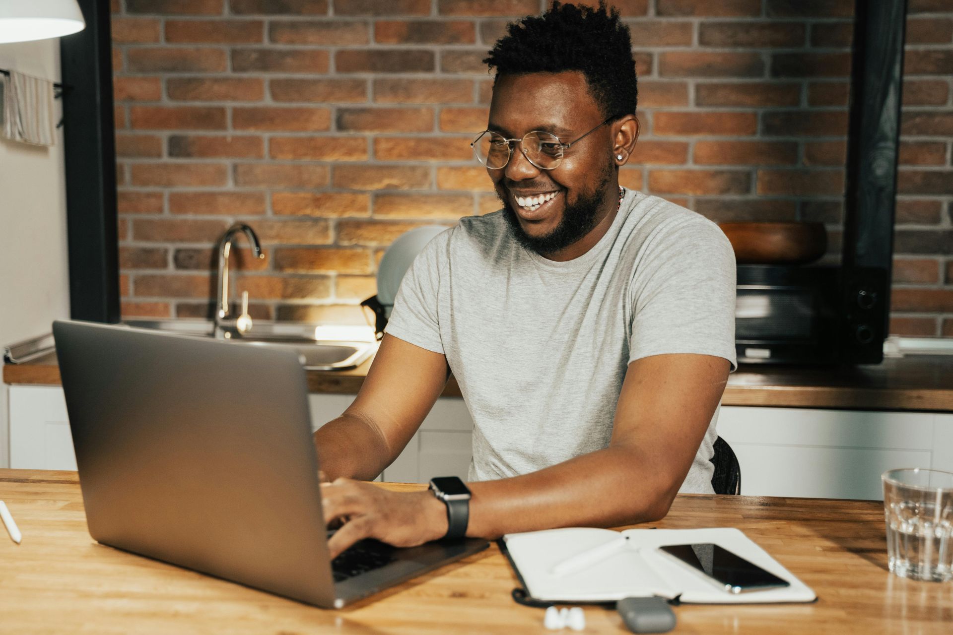 Man smiling while working on a laptop at a wooden table in a kitchen.