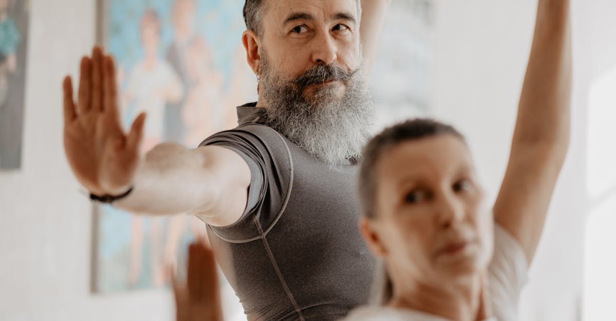 Man with a long gray beard and woman in a yoga pose; indoors with art on the wall.