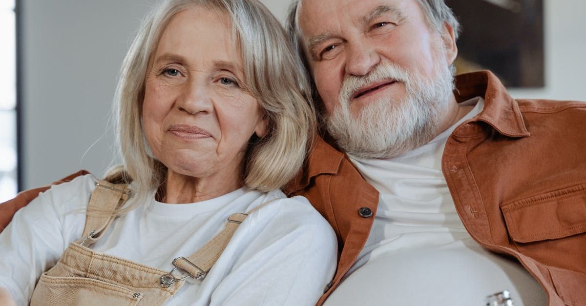 Elderly couple seated together, smiling, indoors. Man in brown shirt, woman in white shirt.