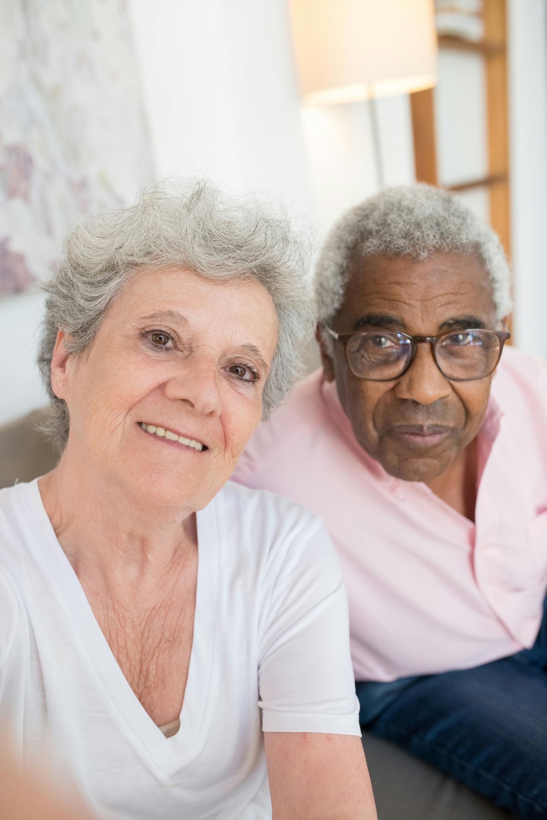 Senior couple, woman smiling, man wearing glasses, seated indoors.