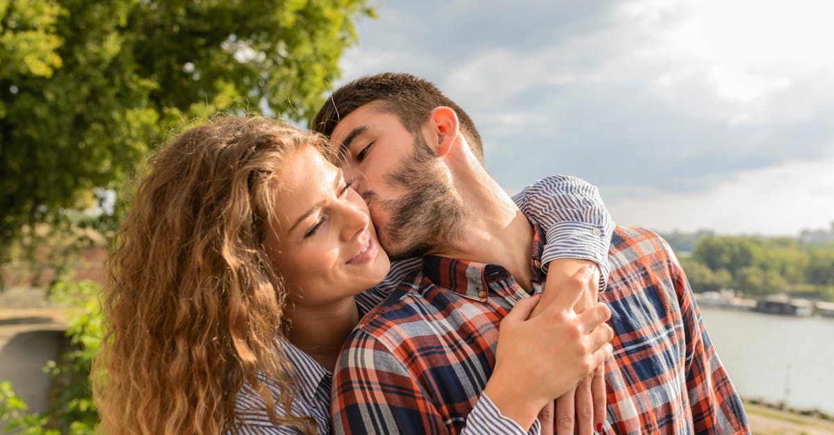 Man kissing a woman's cheek; couple embraces outdoors near a river.