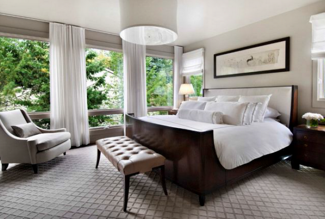 Bedroom with large windows, white curtains, a dark wood bed, light gray chair, and a neutral rug.