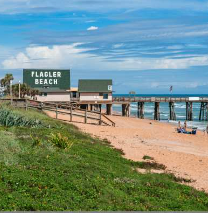 Flagler Beach sign, pier, and lifeguard station on a sandy beach with green vegetation under a blue sky.