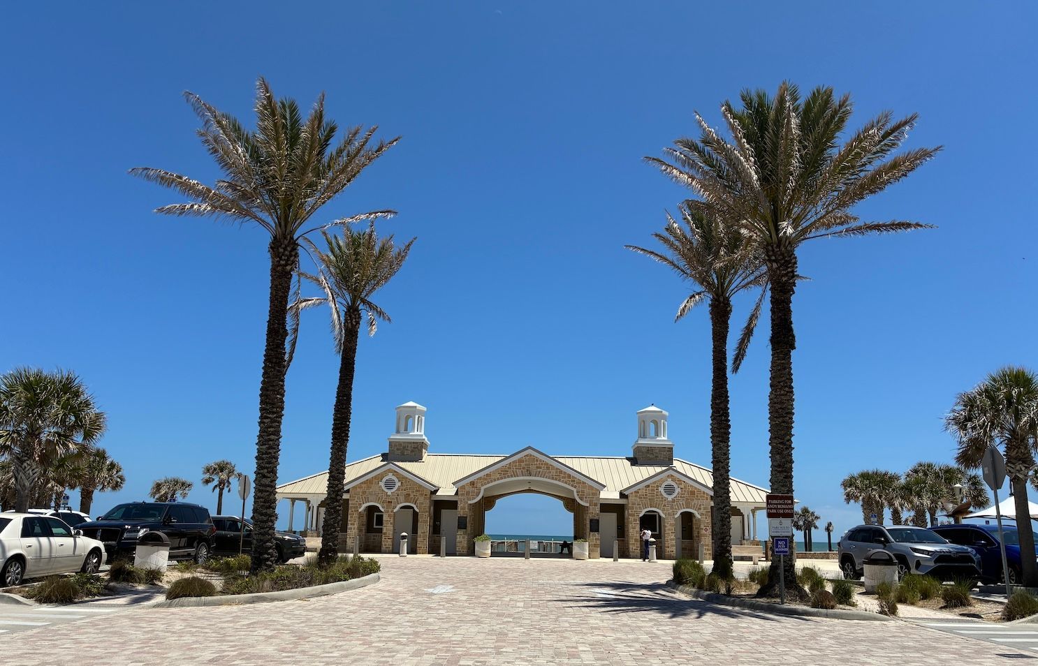 Entrance to beach with palm trees, stone building, blue sky, and parked cars.