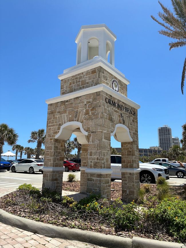 Stone tower with arched openings, clock, and white cupola under a bright blue sky; Ormond Beach sign.
