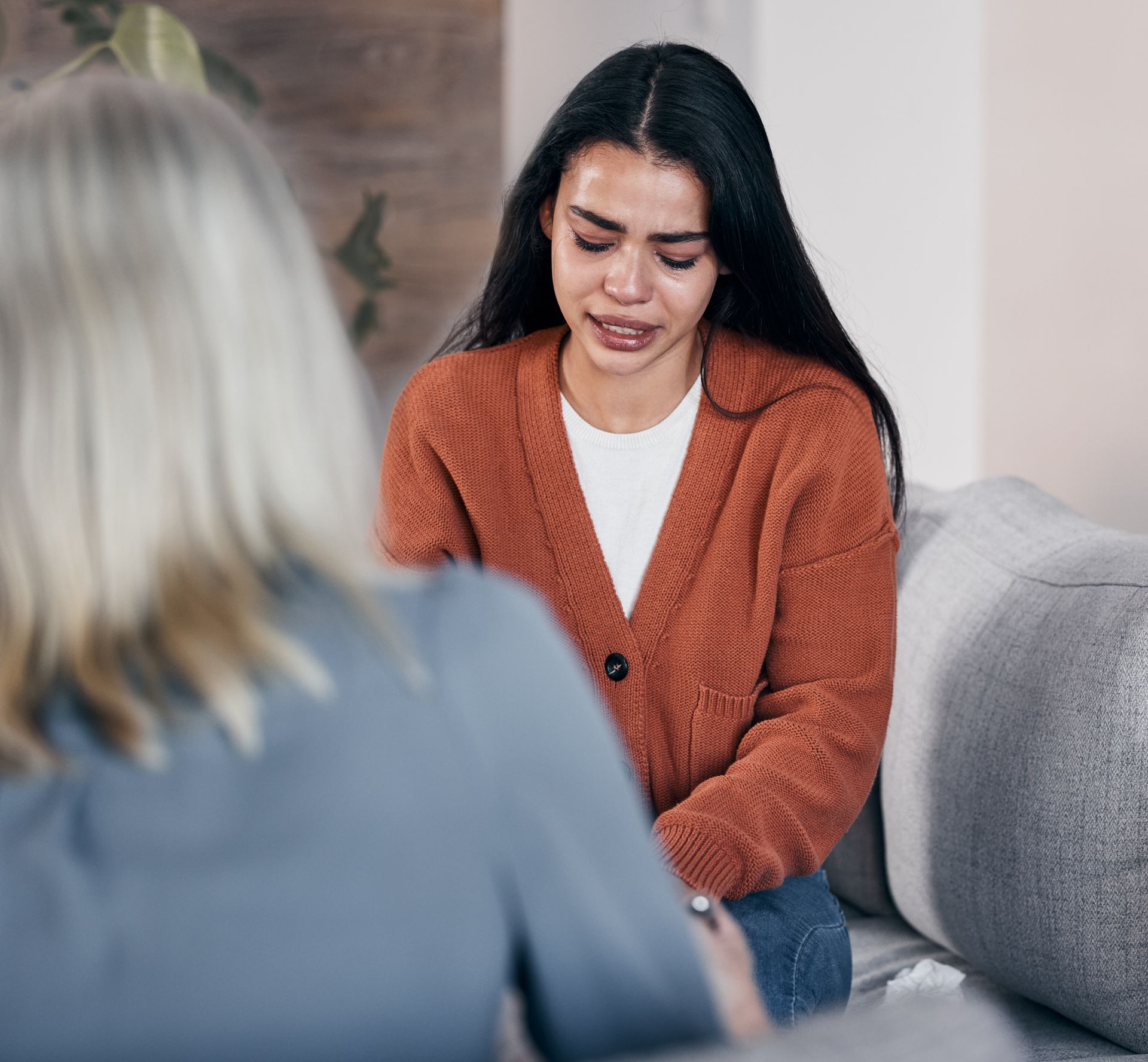 Woman in orange sweater cries during therapy session, sitting on a couch. Another person sits in a chair facing her.