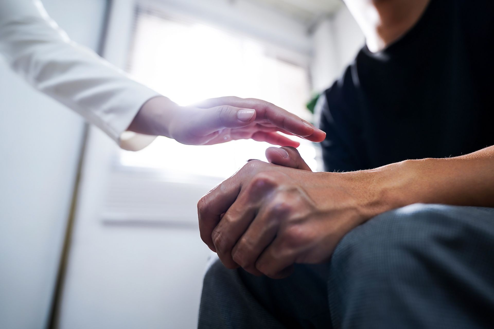 Person's hand is touched by another, offering comfort. Bright room setting.
