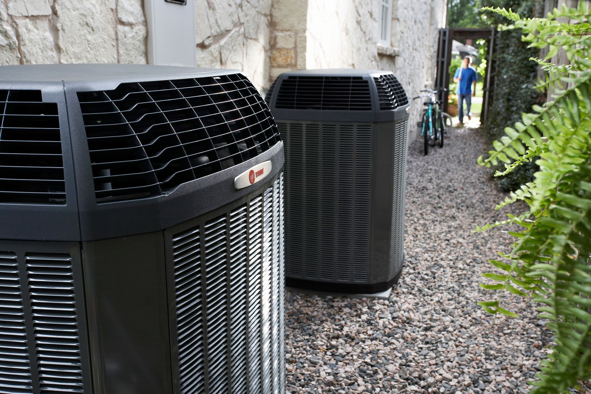 Two large outdoor air conditioning units beside a stone wall and gravel path, with ferns nearby.