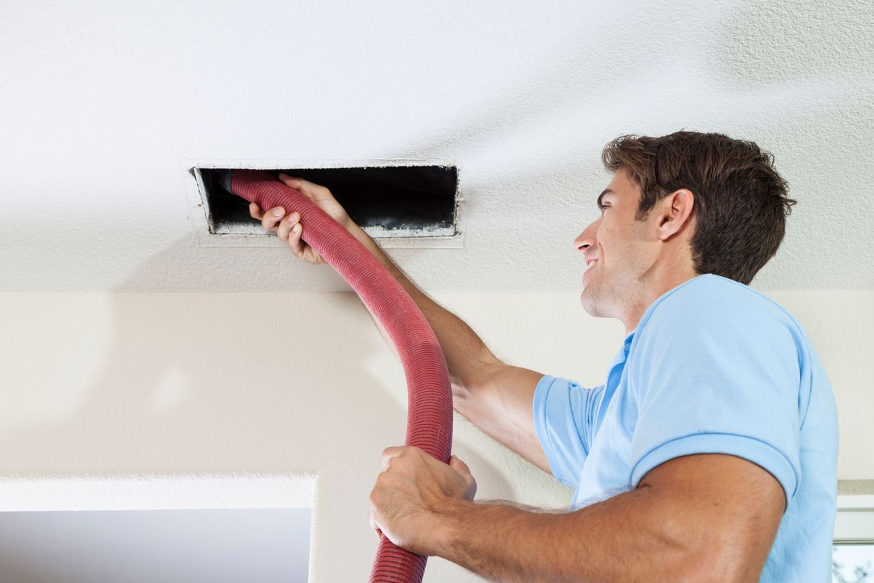 Man cleaning an air vent with a pink vacuum hose on a ceiling