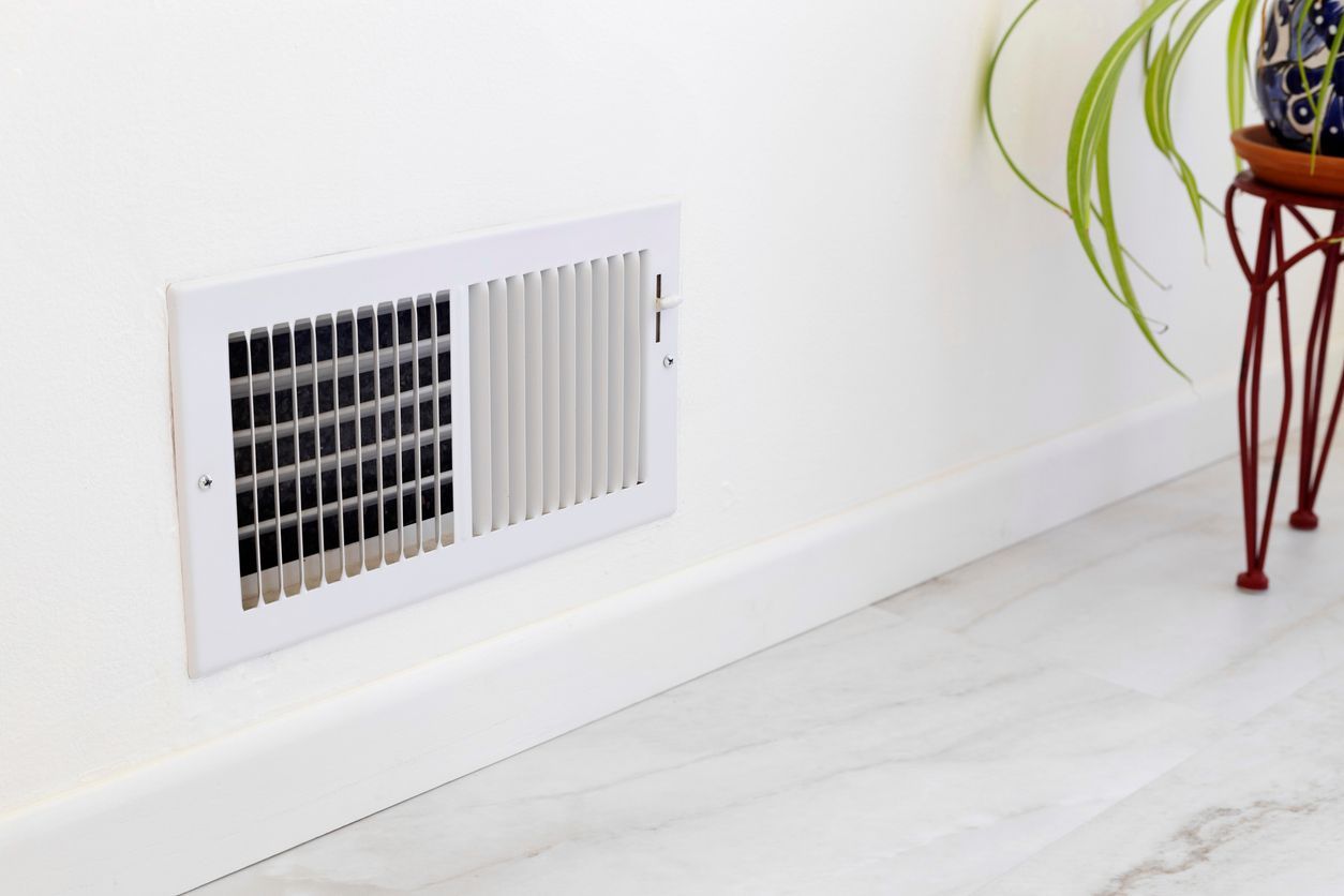 White wall vent near baseboard in a bright room, with a small plant stand on the right.