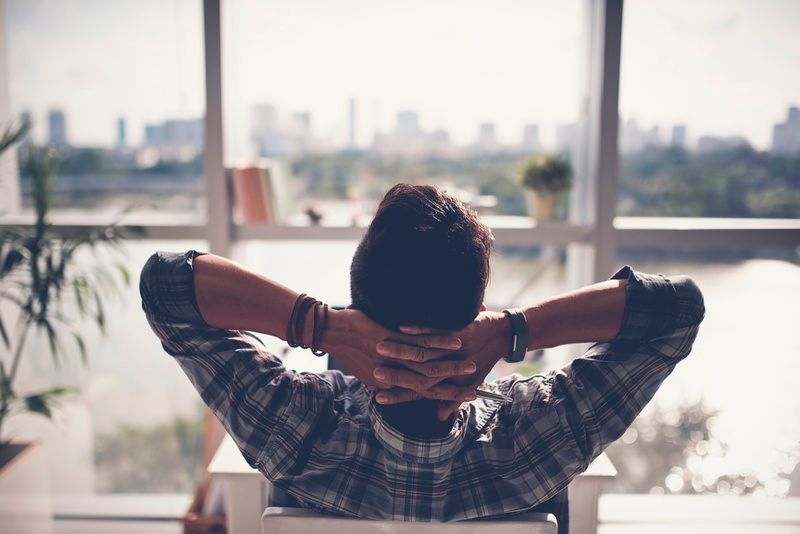 Person relaxing in a chair, hands behind head, looking out a window at a bright city skyline.